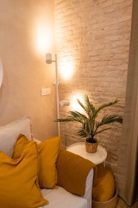 Rincón del salón de Casa Apolo con sofá, cojines mostaza, planta e iluminación cálida sobre pared de ladrillo visto.